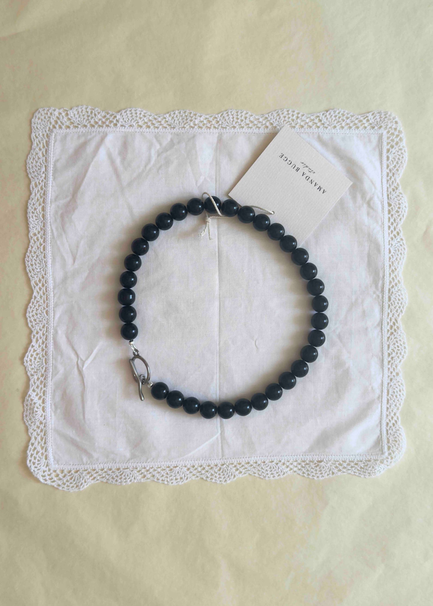 Black beaded necklace with large glossy pearls, styled in a circle on a white lace-edged cloth with product tag from AMANDA BUGGE studio