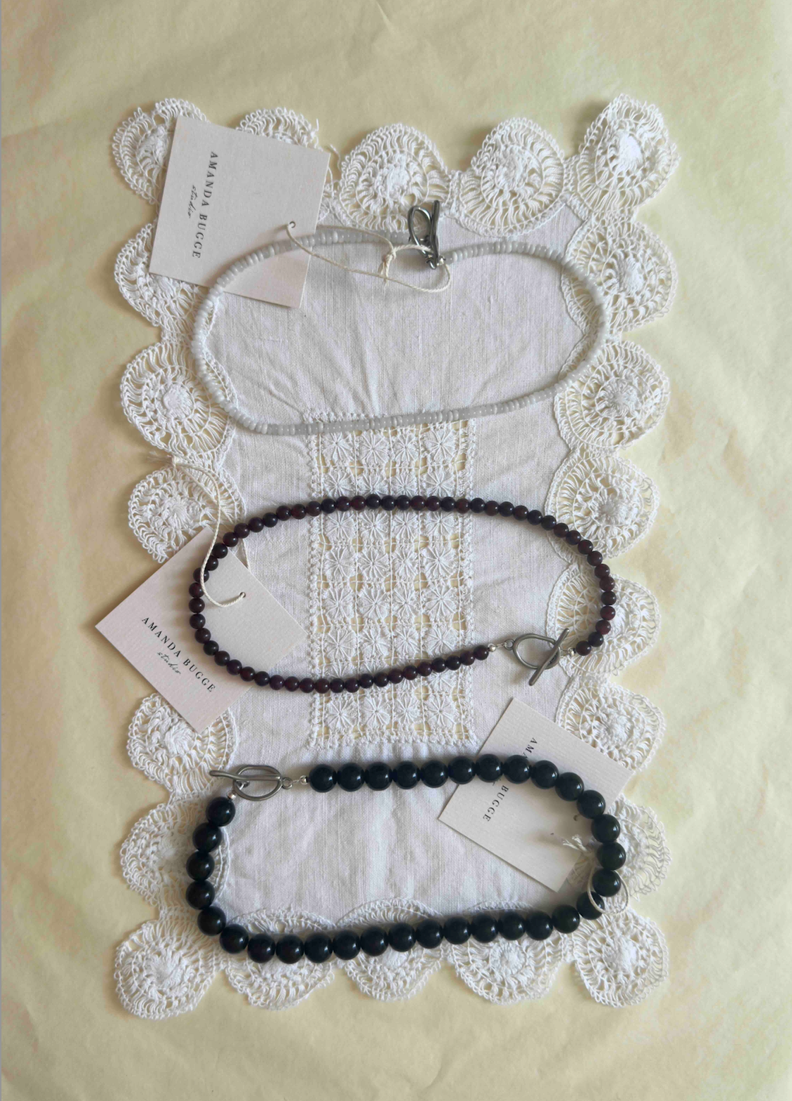 Handmade beaded necklaces by AMANDA BUGGE studio – bold black pearl necklace, petite black beaded necklace, and honey-toned necklace – styled on vintage white lace cloth