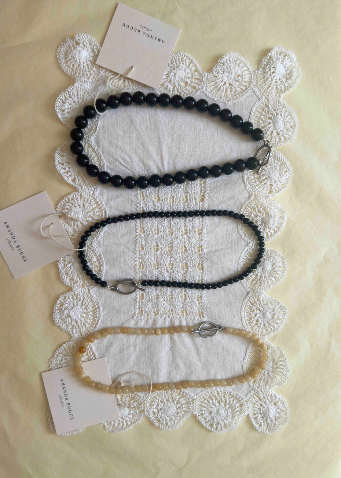 Handmade beaded necklaces by AMANDA BUGGE studio – bold black pearl necklace, petite black beaded necklace, and honey-toned necklace – styled on vintage white lace cloth