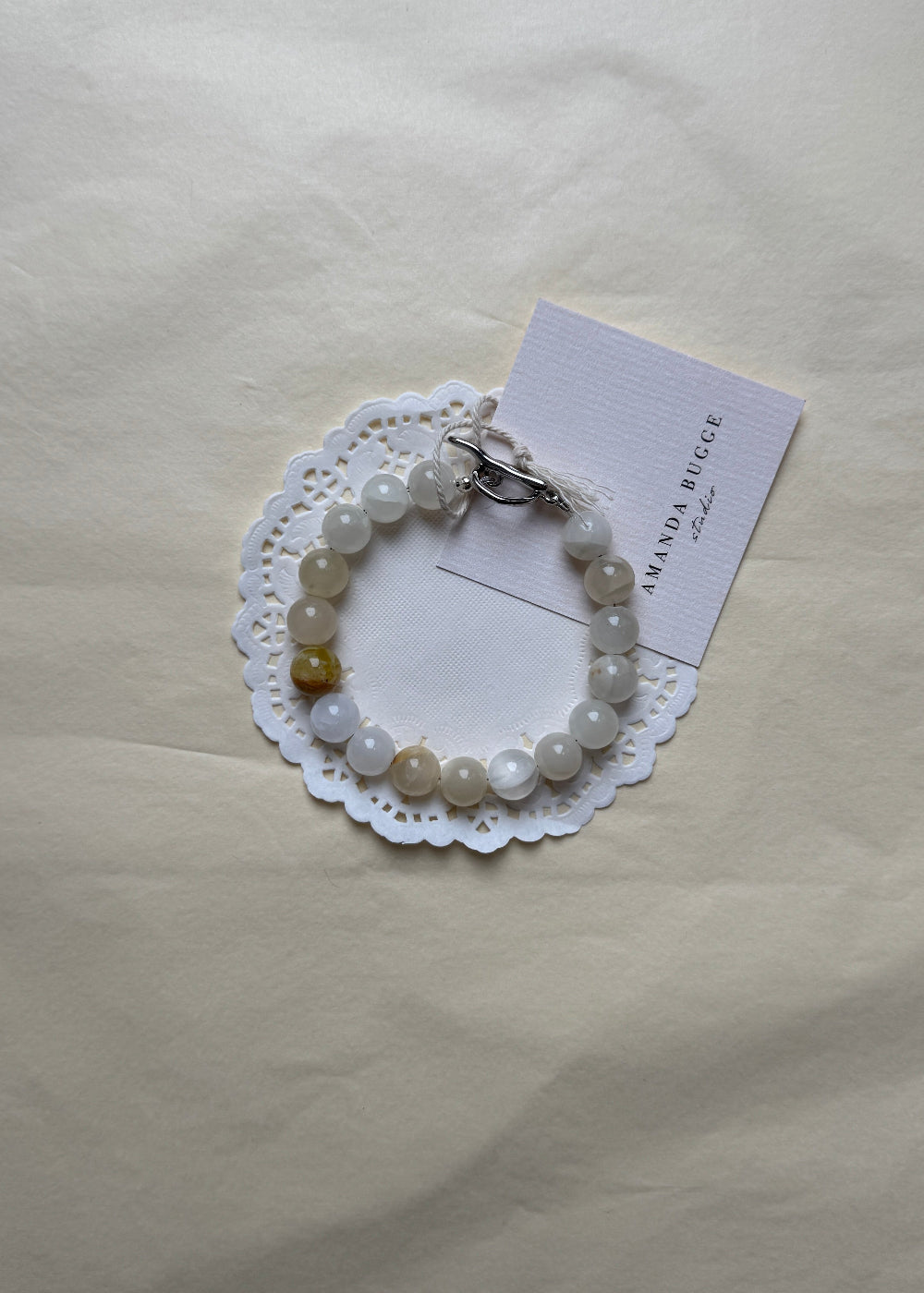 Eggshell beaded bracelet by AMANDA BUGGE studio – handmade jewelry in beige and white tones with silver toggle clasp, displayed on lace-edged cotton cloth