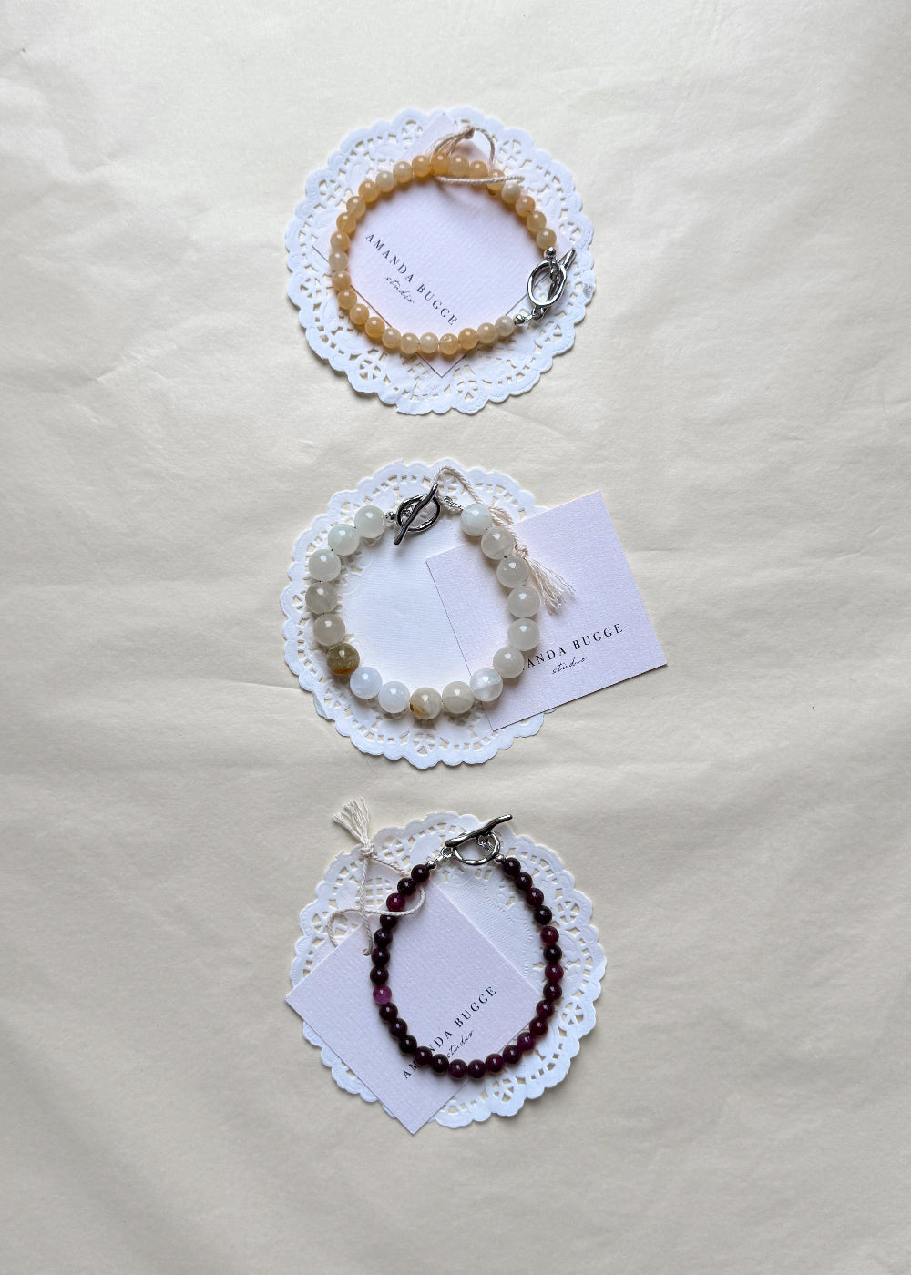 Eggshell, petite honey and petite burgundy beaded bracelet by AMANDA BUGGE studio – handmade jewelry in beige, burgundy, honey and white tones with silver toggle clasp, displayed on lace-edged cotton cloth