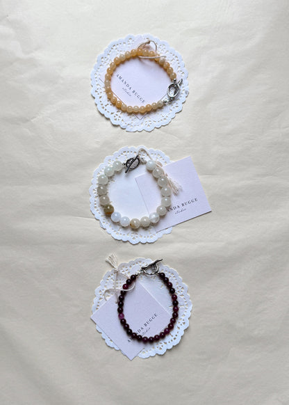 Eggshell, petite honey and petite burgundy beaded bracelet by AMANDA BUGGE studio – handmade jewelry in beige, burgundy, honey and white tones with silver toggle clasp, displayed on lace-edged cotton cloth
