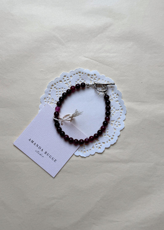 Petite Burgundy beaded bracelet by AMANDA BUGGE studio – handmade jewelry in plum tones with silver toggle clasp, displayed on lace-edged cotton cloth