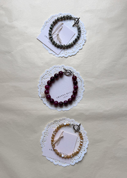 Petite Dalmatian, plum and petite toffee beaded bracelet by AMANDA BUGGE studio – handmade jewelry in dalmatian, plum and beige tones with silver toggle clasp, displayed on lace-edged cotton cloth