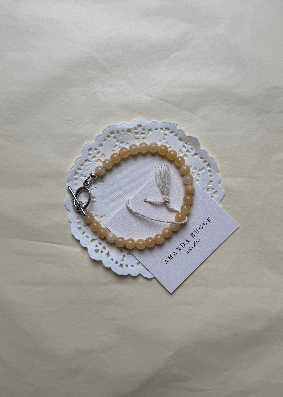 Petite honey beaded bracelet by AMANDA BUGGE studio – handmade jewelry in beige tones with silver toggle clasp, displayed on lace-edged cotton cloth