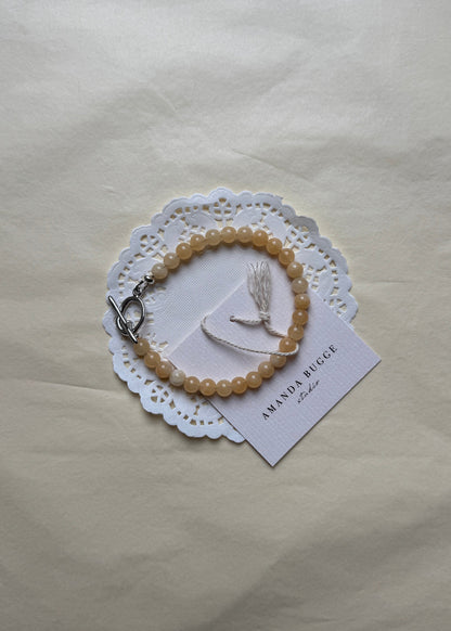 Petite honey beaded bracelet by AMANDA BUGGE studio – handmade jewelry in beige tones with silver toggle clasp, displayed on lace-edged cotton cloth