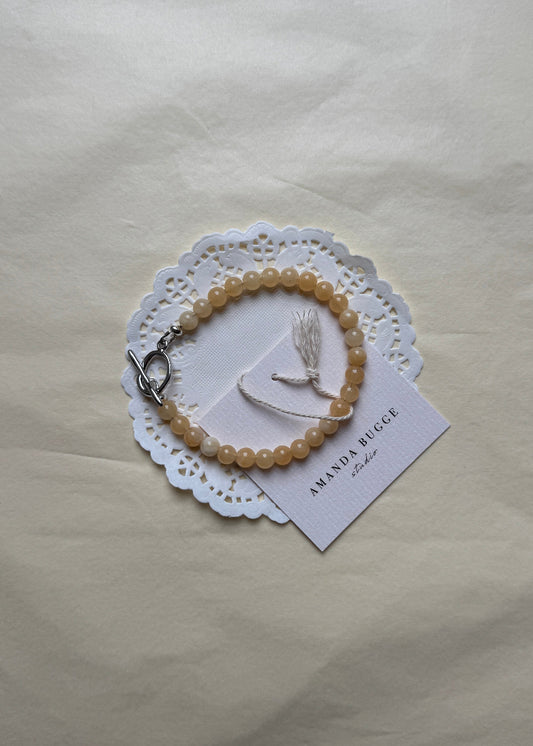 Petite honey beaded bracelet by AMANDA BUGGE studio – handmade jewelry in beige tones with silver toggle clasp, displayed on lace-edged cotton cloth