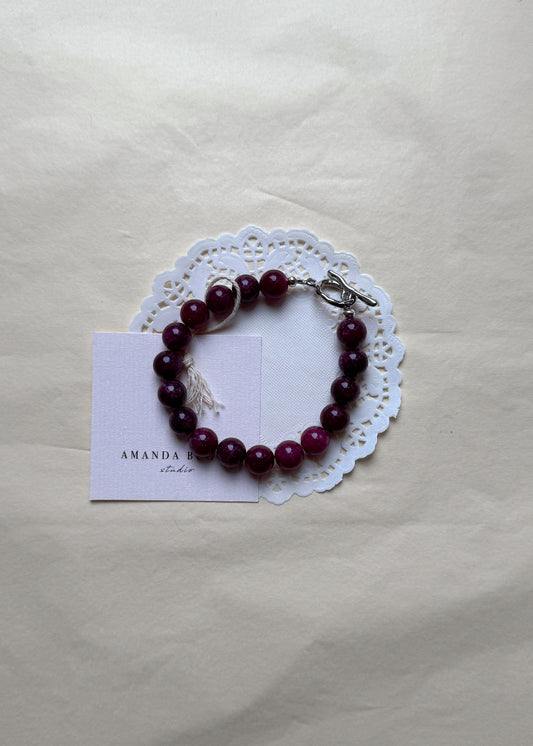 Plum beaded bracelet by AMANDA BUGGE studio – handmade jewelry in plum tones with silver toggle clasp, displayed on lace-edged cotton cloth