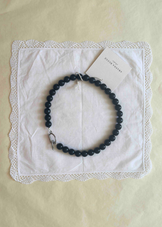 Black beaded necklace with large glossy pearls, styled in a circle on a white lace-edged cloth with product tag from AMANDA BUGGE studio