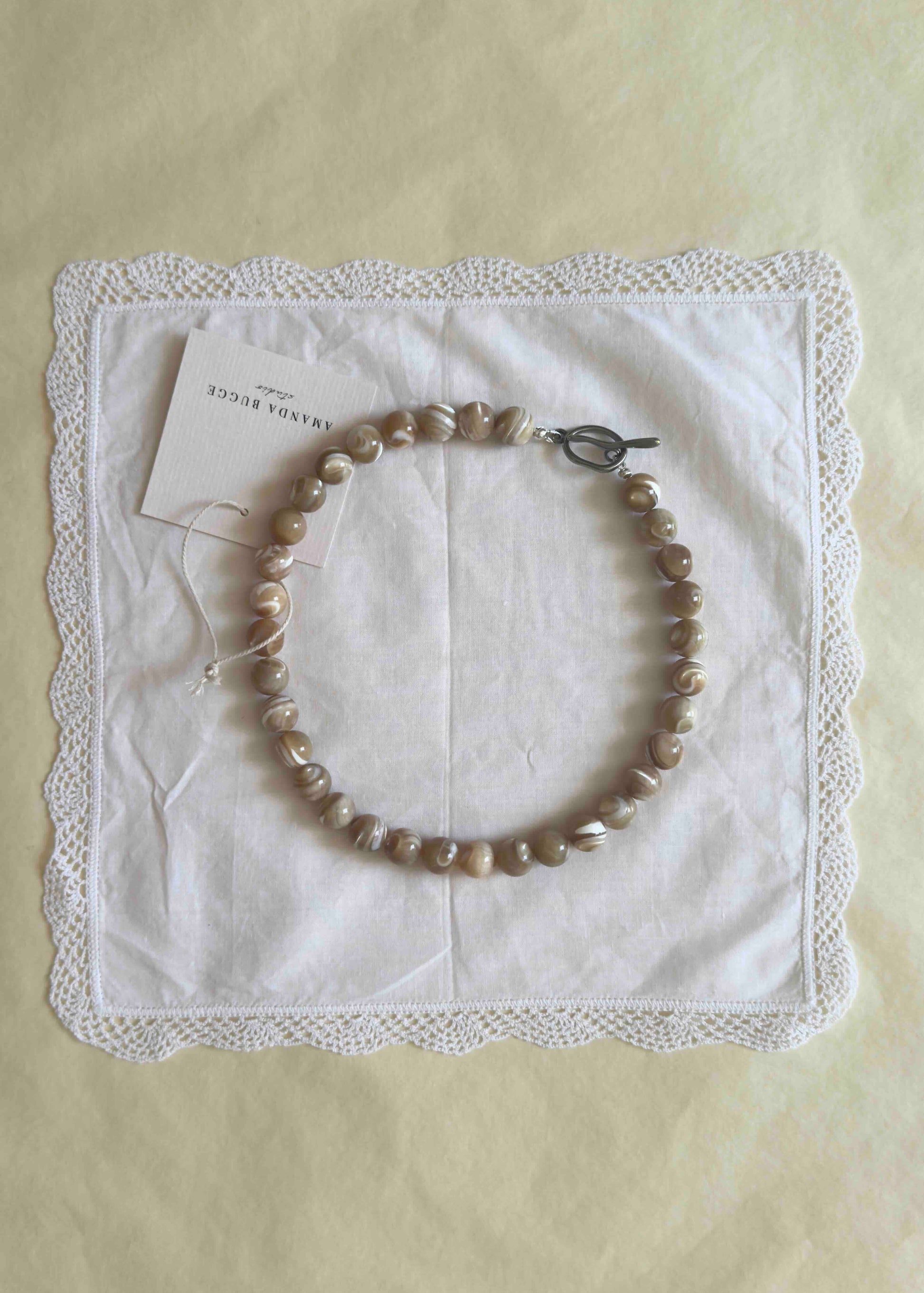 Black beaded necklace with large glossy pearls, styled in a circle on a white lace-edged cloth with product tag from AMANDA BUGGE studio