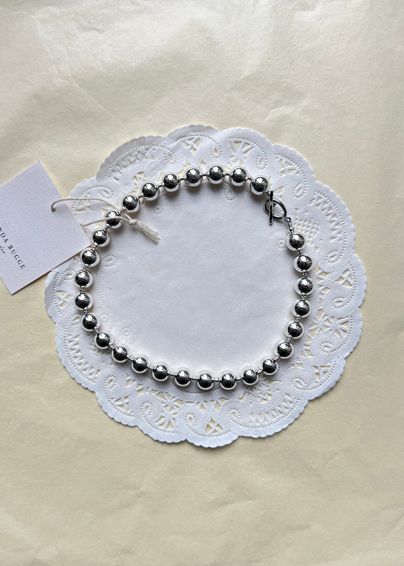 Bold silver necklace by AMANDA BUGGE studio – minimalist silver beaded choker necklace with silver toggle clasp, styled on white lace-edged lace fabric