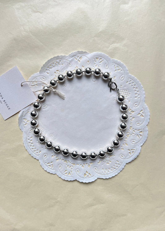 Bold silver necklace by AMANDA BUGGE studio – minimalist silver beaded choker necklace with silver toggle clasp, styled on white lace-edged lace fabric